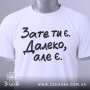Футболка зате ти є. Білий колір, зображена зверху, складена на поверхні, фон світлий, принт чорного кольору з текстом 'Зате ти є. Далеко, але є.', з етикеткою зеленого кольору.