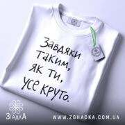 Футболка завдяки таким людям, білого кольору, складена на поверхні, фон світло-сірий, принт чорного кольору з текстом 'Завдяки таким, як ти, усе круто.', на комірці видна етикетка з зеленим шнурком.