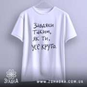 Футболка завдяки таким людям, білого кольору, підвішена на вішалці, фон світло-сірий, принт чорного кольору з текстом 'Завдяки таким, як ти, усе круто.'