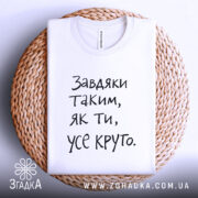 Футболка завдяки таким людям, білого кольору, складена на кошику з плетеного матеріалу, фон світло-сірий, принт чорного кольору з текстом 'Завдяки таким, як ти, усе круто.'