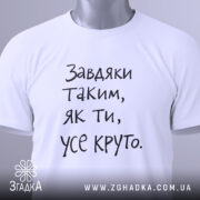 Футболка завдяки таким людям, білого кольору, складена на поверхні, фон світло-сірий, принт чорного кольору з текстом 'Завдяки таким, як ти, усе круто.', на комірці видна етикетка з зеленим шнурком.