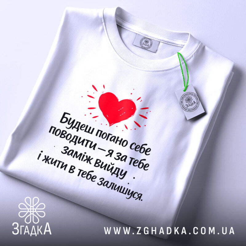 Футболка жарт про шлюб, біла, зображена зверху, фон світлий, принт червоного серця з текстом чорного кольору, на комірці є етикетка.