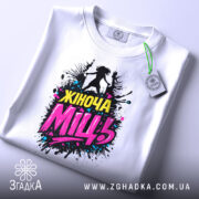 Футболка жіноча міць, білого кольору, складена на поверхні, фон світлий, принт з написом 'Жіноча Міць' у яскравих кольорах, з елементами графіки, на футболці є етикетка з зеленим шнурком.