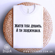Футболка життєвий тиск, біла, складена на плетеній кошику, фон світло-сірий, принт чорного кольору з текстом 'Життя тебе душить. А ти збуджуєшся.'