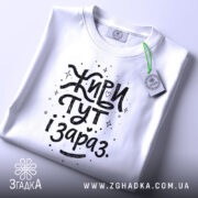 Футболка живи тут і зараз. Біла футболка, складена на столі, з видимим етикеткою, фон світло-сірий, принт чорного кольору з написом 'Живи тут і зараз' у веселому стилі.