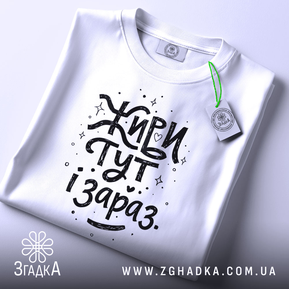 Футболка живи тут і зараз. Біла футболка, складена на столі, з видимим етикеткою, фон світло-сірий, принт чорного кольору з написом 'Живи тут і зараз' у веселому стилі.