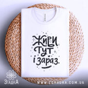 Футболка живи тут і зараз. Біла футболка, складена на плетеній основі, фон світло-сірий, принт чорного кольору з написом 'Живи тут і зараз' у креативному стилі.