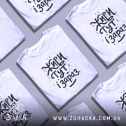 Футболка живи тут і зараз. Біла футболка з коротким рукавом, зображена на манекені, фон світло-сірий, принт чорного кольору з написом 'Живи тут і зараз' у креативному стилі з зірочками та сердечками.