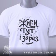 Футболка живи тут і зараз. Біла футболка, складена на столі, з видимим етикеткою, фон світло-сірий, принт чорного кольору з написом 'Живи тут і зараз' у веселому стилі.