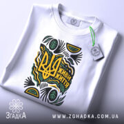 Футболка живи життя білого кольору, складена на поверхні, з принтом у вигляді тризуба та написом 'Живи життя', прикрашена зеленими та жовтими елементами, з етикеткою, що звисає з коміра.