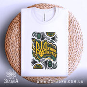 Футболка живи життя білого кольору, складена на плетеній кошику, з принтом у вигляді тризуба та написом 'Живи життя', прикрашена зеленими та жовтими елементами, фон світлий.