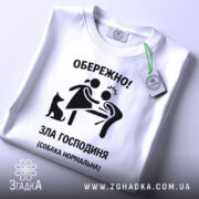 Футболка зла господиня, біла, зображена зверху, фон світлий, принт чорного кольору з малюнком жінки та чоловіка, а також чорна собака, на футболці є етикетка з зеленим шнурком.