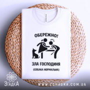 Футболка зла господиня, біла, зображена зверху на плетеному кошику, фон світлий, принт чорного кольору з малюнком жінки та чоловіка, а також чорна собака.