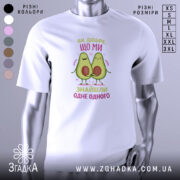 Футболка знайшли одне одного. Біла футболка з коротким рукавом, зображена на манекені, фон світло-сірий. Принт з двома авокадо, які тримаються за руки, з написом 'Як добре, що ми знайшли одне одного' у кольорах рожевого та зеленого.