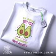 Футболка знайшли одне одного. Біла футболка, складена на столі, з видимим етикеткою. Принт з двома авокадо, які тримаються за руки, з написом 'Як добре, що ми знайшли одне одного' у кольорах рожевого та зеленого.