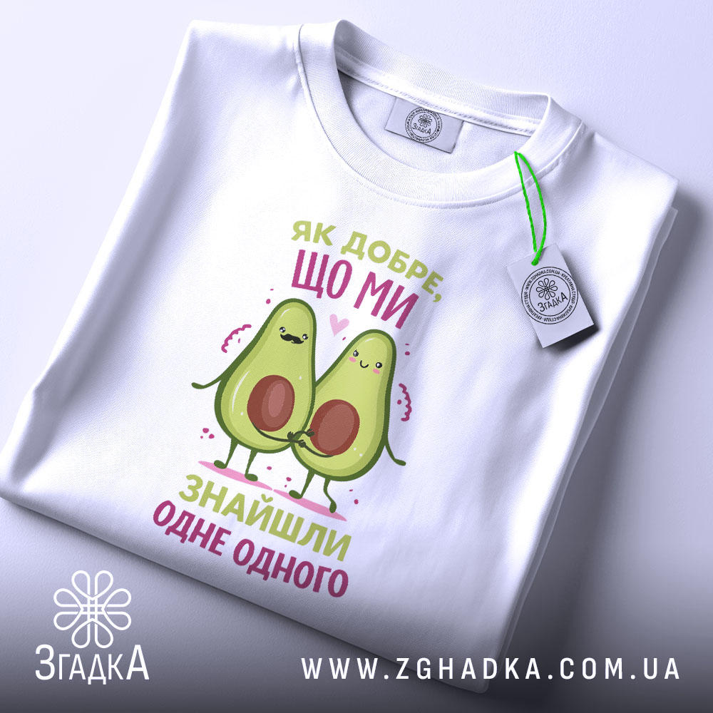 Футболка знайшли одне одного. Біла футболка, складена на столі, з видимим етикеткою. Принт з двома авокадо, які тримаються за руки, з написом 'Як добре, що ми знайшли одне одного' у кольорах рожевого та зеленого.