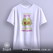 Футболка знайшли одне одного. Біла футболка з коротким рукавом, підвішена на вішалці, фон світло-сірий. Принт з двома авокадо, які тримаються за руки, з написом 'Як добре, що ми знайшли одне одного' у кольорах рожевого та зеленого.