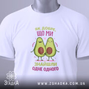 Футболка знайшли одне одного. Біла футболка, складена на столі, з видимим етикеткою. Принт з двома авокадо, які тримаються за руки, з написом 'Як добре, що ми знайшли одне одного' у кольорах рожевого та зеленого.