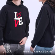 Худі для закоханих, темно-синього кольору, з капюшоном, на якому видно пару моделей, які носять худі. Принт 'LOVE' яскравий, з сердечком на букві 'O'. Моделі стоять на світлому фоні, що підкреслює стиль та комфорт худі.