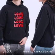 Худі Love Yourself чорного кольору, ракурс з боку, фон світлий, принт з написом 'LOVE Yourself' червоного кольору, капюшон видимий, чоловік та жінка в худі, кишеня-«кенгуру» спереду, манжети та пояс з еластичної тканини.