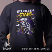 Світшот Бля яке я старе, чорного кольору, з ракурсу спереду, фон розмитий, принт з скелетом у капюшоні, червоні і жовті очі, кругла горловина, манжети на рукавах, трикотажна резинка на поясі, фактура м'яка і комфортна.