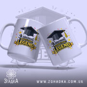 Чашка для легендарного класного керівника, дві білі чашки з глянцевою поверхнею, одна з них повернена до камери, на обох зображення з чорним капелюхом випускника та написом 'Класний керівник рівня: LEGEND', фон світло-фіолетовий з елементами зірок.