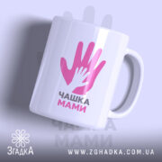 Чашка для мами, біла, з ракурсом зліва, на фоні світло-фіолетового кольору, з принтом у вигляді двох рук, одна з яких менша, напис "ЧАШКА МАМИ" чорним шрифтом, ручка кругла, край рівний, глянцевий.