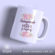 Чашка для найкращої няні, біла, з ракурсу спереду, на фоні ніжно-фіолетового кольору, прикрашена квітковим принтом і написом "Найкращий НЯНІ у садочку", ручка округлої форми, край рівний, глянцева поверхня.