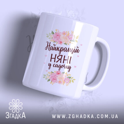 Чашка для найкращої няні, біла, з ракурсу спереду, на фоні ніжно-фіолетового кольору, прикрашена квітковим принтом і написом "Найкращий НЯНІ у садочку", ручка округлої форми, край рівний, глянцева поверхня.