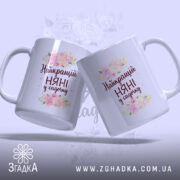 Чашка для найкращої няні, біла, з ракурсу збоку, на фоні ніжно-фіолетового кольору, дві чашки з написом "Найкращий НЯНІ у садочку" та квітковим принтом, ручки округлі, краї рівні, глянцева поверхня.