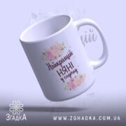 Чашка для найкращої няні, біла, з ракурсу зверху, на фоні ніжно-фіолетового кольору, з написом "Найкращий НЯНІ у садочку" та квітковим принтом, ручка округлої форми, край рівний, глянцева поверхня.