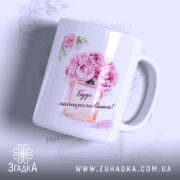 Чашка для подарунку на свято, біла з яскравим принтом, зображення квітів і написом "Будь найщасливішою!", ручка класична, край рівний, глянцеве покриття, фон світлий з легким малюнком квітів.