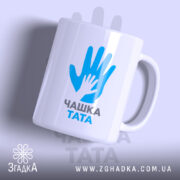 Чашка для тата, біла, зображена з легким нахилом, на фоні світло-фіолетового кольору, з принтом у вигляді рук блакитного кольору та написом 'ЧАШКА ТАТА' чорного кольору, ручка округлої форми, край рівний, глянцева поверхня.