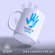 Чашка для тата, біла, зображена з легким нахилом, на фоні світло-фіолетового кольору, з принтом у вигляді рук блакитного кольору та написом 'ЧАШКА ТАТА' чорного кольору, ручка округлої форми, край рівний, глянцева поверхня.