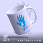 Чашка для тата, біла, зображена з легким нахилом, на фоні світло-фіолетового кольору, з принтом у вигляді рук блакитного кольору та написом 'ЧАШКА ТАТА' чорного кольору, ручка округлої форми, край рівний, глянцева поверхня.