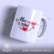 Чашка Моє серце – твоє, біла, з легким глянцем, з написом чорного та червоного кольорів, ручка з традиційним дизайном, край рівний, фон ніжно-фіолетовий, на задньому плані розмитий текст.
