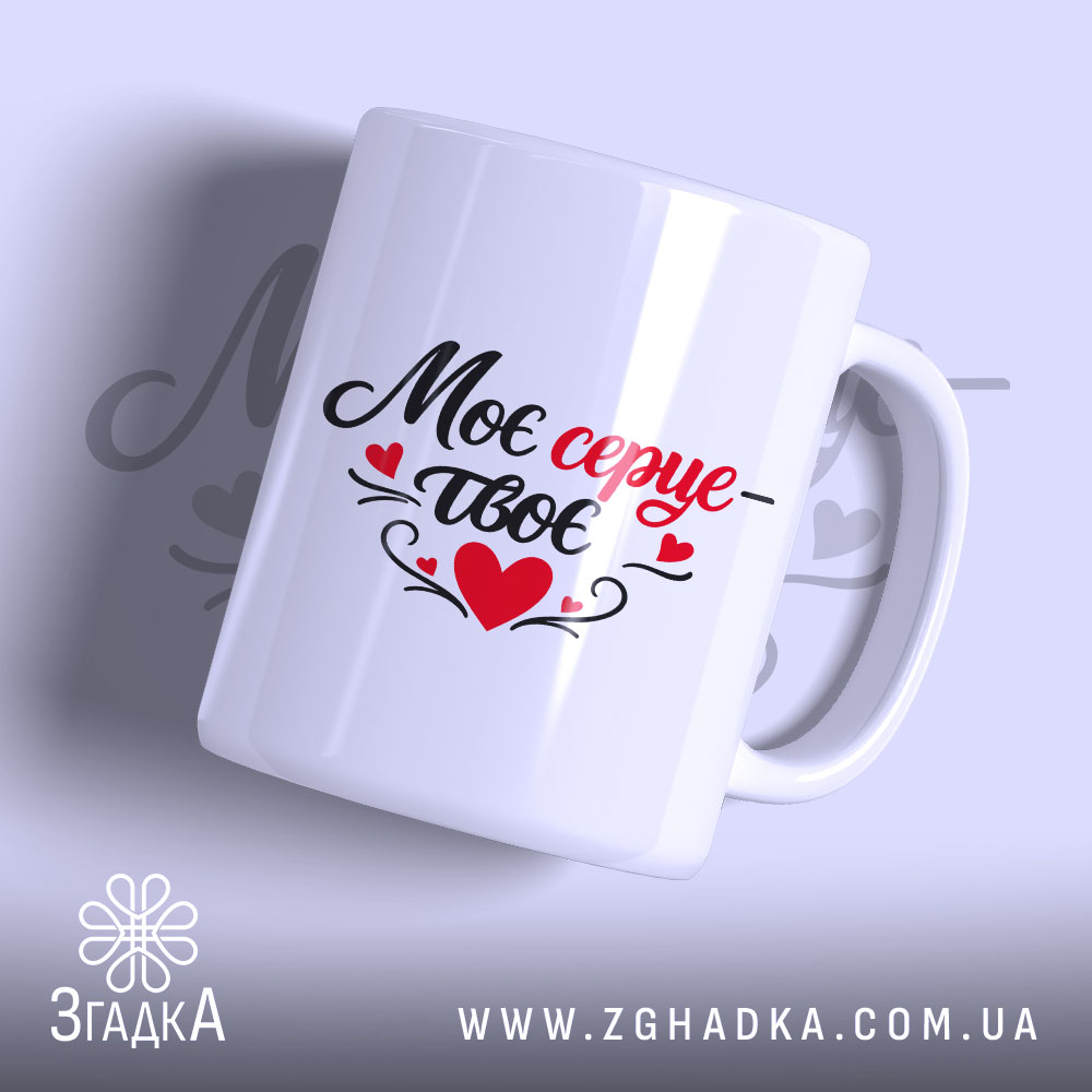 Чашка Моє серце – твоє, біла, з легким глянцем, з написом чорного та червоного кольорів, ручка з традиційним дизайном, край рівний, фон ніжно-фіолетовий, на задньому плані розмитий текст.