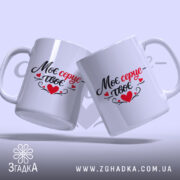 Чашка Моє серце – твоє, дві білих чашки з легким глянцем, розташовані під кутом, з написом чорного та червоного кольорів, ручки традиційної форми, фон ніжно-фіолетовий, на задньому плані розмитий текст.