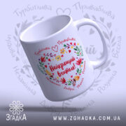 Чашка Найкращий Вчитель, біла з кольоровим принтом, показана з верхнього ракурсу, на фоні світло-фіолетового кольору, з написом "Найкращий вчитель!" в центрі, оточеним квітами і сердечками, ручка класична, край рівний, глянцевий.