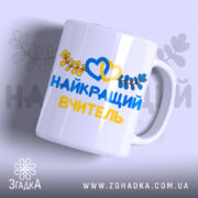 Чашка патріотична для вчителя, біла, з написом 'НАЙКРАЩИЙ ВЧИТЕЛЬ' жовтого та синього кольорів, оформлена зображенням сердець та листя, ручка округлої форми, край гладкий, глянцева поверхня, фон світло-фіолетовий.