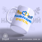 Чашка патріотична для вчителя, біла, з написом 'НАЙКРАЩИЙ ВЧИТЕЛЬ' жовтого та синього кольорів, оформлена зображенням сердець та листя, ручка округлої форми, край гладкий, глянцева поверхня, фон світло-фіолетовий.
