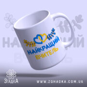 Чашка патріотична для вчителя, біла, з написом 'НАЙКРАЩИЙ ВЧИТЕЛЬ' жовтого та синього кольорів, оформлена зображенням сердець та листя, ручка округлої форми, край гладкий, глянцева поверхня, фон світло-фіолетовий.