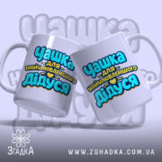 Чашка подарунок найкращому дідусю. Дві чашки білого кольору, розташовані під кутом, з принтом 'Чашка для найулюбленішого Дідуся'. Ракурс зліва, фон світло-фіолетовий, ручки округлі, краї гладкі, поверхня глянцева.