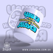 Чашка подарунок найкращому дідусю. Чашка білого кольору з принтом 'Чашка для найулюбленішого Дідуся', зображена знизу. Ракурс з правого боку, фон світло-фіолетовий, ручка округлої форми, край гладкий, поверхня глянцева.