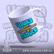 Чашка подарунок найкращому дідусю. Чашка білого кольору з принтом 'Чашка для найулюбленішого Дідуся', зображена з верхнього ракурсу. Фон світло-фіолетовий, ручка округлої форми, край гладкий, поверхня глянцева.