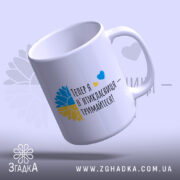 Чашка привітання найкращому вчителю, біла з кольоровим принтом, зображення соняшника в жовто-блакитних кольорах, напис чорним шрифтом, ручка класична, край рівний, глянцеве покриття, чашка нахилена.