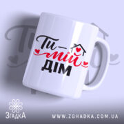 Чашка Ти – мій дім, біла з чорним і червоним принтом, з написом 'Ти – мій дім', ручка з класичним дизайном, край рівний, матова поверхня, фон світло-фіолетовий.