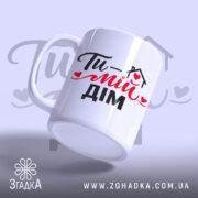 Чашка Ти – мій дім, біла з чорним і червоним принтом, з написом 'Ти – мій дім', ручка з класичним дизайном, край рівний, матова поверхня, фон світло-фіолетовий.