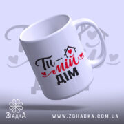 Чашка Ти – мій дім, біла з чорним і червоним принтом, з написом 'Ти – мій дім', ручка з класичним дизайном, край рівний, матова поверхня, фон світло-фіолетовий.