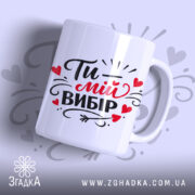Чашка Ти – мій вибір, біла з яскравим чорним і червоним принтом, показана з бокового ракурсу на світло-сірому фоні, з написом "Ти – мій вибір" та декоративними елементами у вигляді сердечок, має гладку ручку та рівний край, з глянцевою поверхнею.